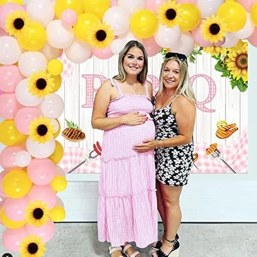 a pregnant women standing in front of a pink balloon arch with sunflowers