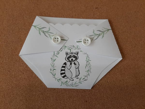 A white diaper shaped gift card holder with a woodland raccoon on it