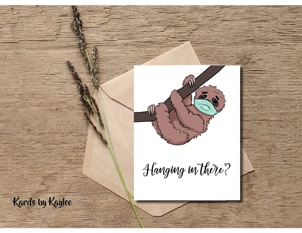 card with sloth hanging from a branch with a mask on says "hanging in there?"